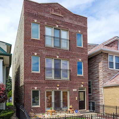 A three-story red brick apartment building with balconies, large windows, and a fenced front yard on a residential street under a partly cloudy sky.