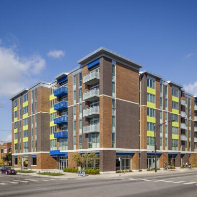A modern five-story apartment building with brick and colorful panels, featuring balconies and retail space on the ground floor, located at a street corner under a clear sky.