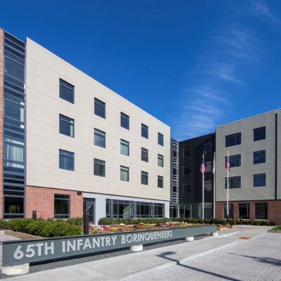 A modern four-story building with a brick and beige facade, displaying a sign that reads "65th Infantry Borinqueneers" in front, with three flagpoles nearby.