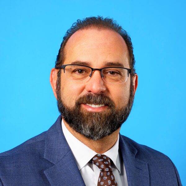 A man with glasses and a beard, wearing a blue suit, white shirt, and patterned tie, poses in front of a plain blue background.