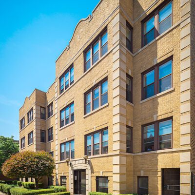 Four-story yellow brick apartment building with multiple windows, landscaped bushes, and a sidewalk under a clear blue sky.