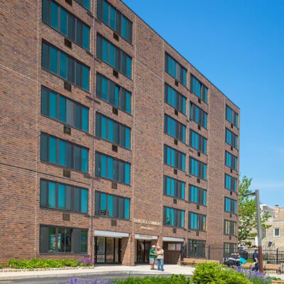 A six-story brick apartment building with large windows, a main entrance canopy, landscaped flower beds, and people walking near the entrance on a sunny day.