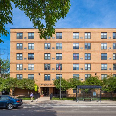 Four-story brick apartment building with an entrance sign reading "Las Moradas," trees in front, a bus stop shelter, and cars parked along the street.