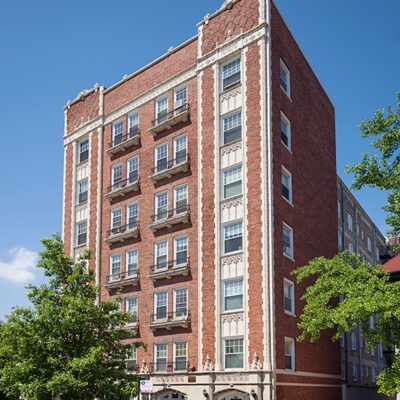 A six-story red brick apartment building with decorative stone details, small balconies, and a gated entrance, set against a clear blue sky.