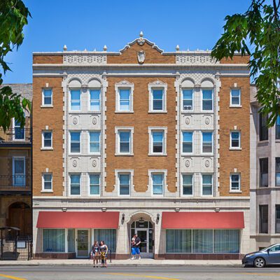 A four-story brick apartment building with ornate stone details, red awnings over ground-floor windows, and people standing on the sidewalk in front.