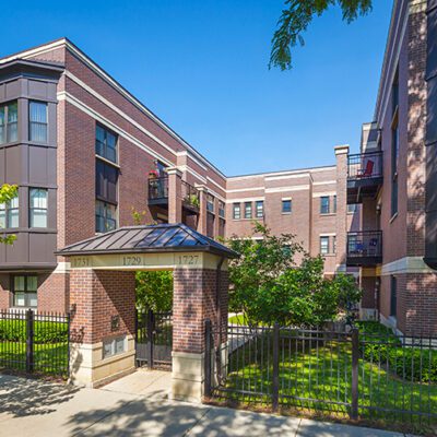 Three-story brick apartment buildings with black railings and a gated entrance, surrounded by greenery and trees under a clear blue sky.