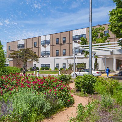 A three-story building with a garden in front, featuring colorful flowers, trees, and a walkway leading to the main entrance.