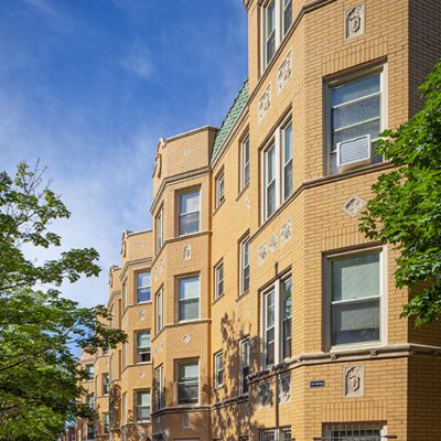 Four-story yellow brick apartment building with bay windows and decorative stonework, set along a tree-lined sidewalk under a blue sky.