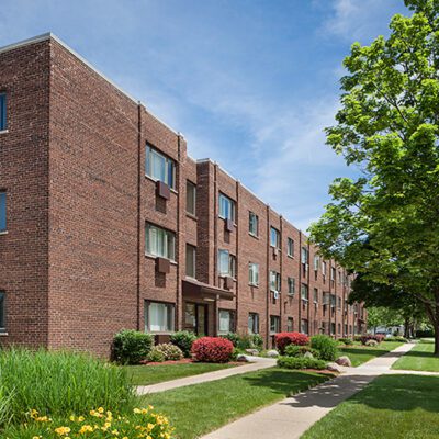 Three-story brick apartment building with multiple windows, surrounded by green grass, shrubs, trees, and a sidewalk on a sunny day.