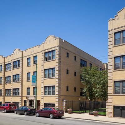 A three-story tan brick apartment building with decorative rooflines, large windows, and several parked cars along the street under a clear blue sky.