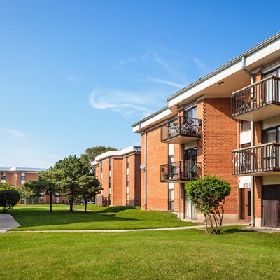 Red-brick apartment buildings with balconies surround a well-kept green lawn under a clear blue sky.