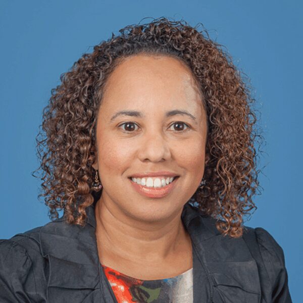 A woman with curly hair smiles at the camera against a blue background, wearing a dark jacket and a colorful top.