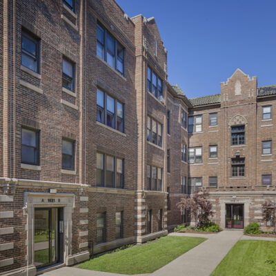 A four-story brick apartment building surrounds a rectangular courtyard with grass, shrubs, and concrete walkways under a clear blue sky.