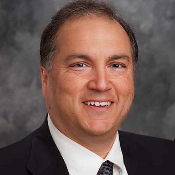 A man in a suit and tie smiles at the camera against a gray, mottled background.
