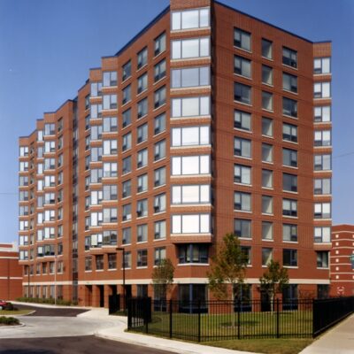 A multi-story brick apartment building with large windows, surrounded by a black metal fence and small trees, on a sunny day.