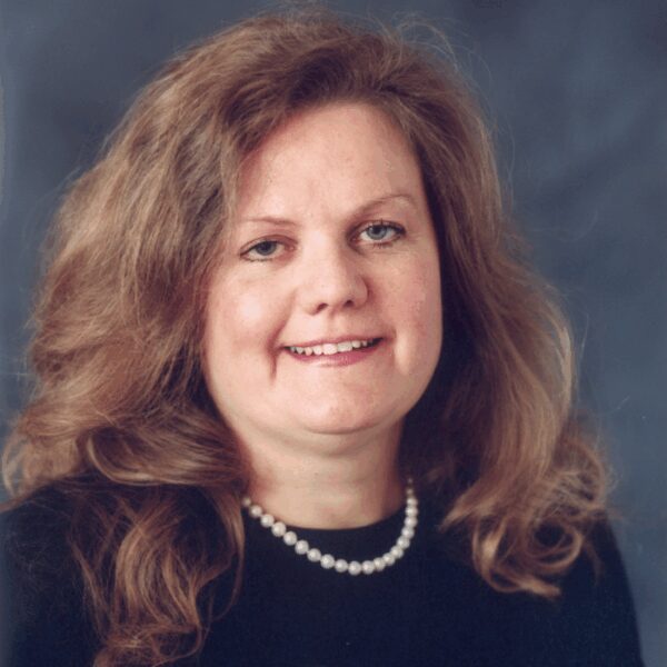 A woman with long, wavy light brown hair is wearing a black top and a pearl necklace, posing in front of a plain, dark background.