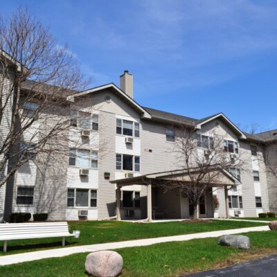 Three-story apartment building with beige siding, multiple windows, and a covered entrance; trees, a bench, grass, and rocks in the landscaped front yard.