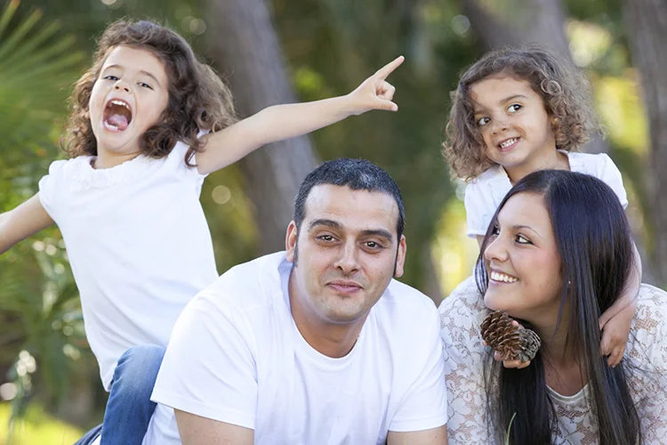 A family of four poses outdoors; a man and woman sit close while two young children, both with curly hair, smile and play behind them. All are wearing white shirts.