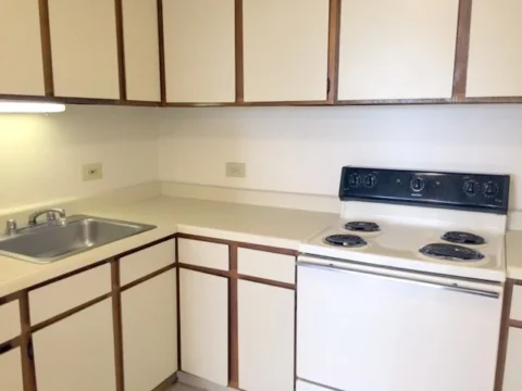 A white kitchen with a 4-burner stove, overhead and under counter cabinets, and metal sink.