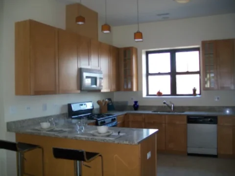The kitchen of a Boulevard Court apartment, showing overhead wooden cabinets, a large window over a double sink, and a bar with two barstools.