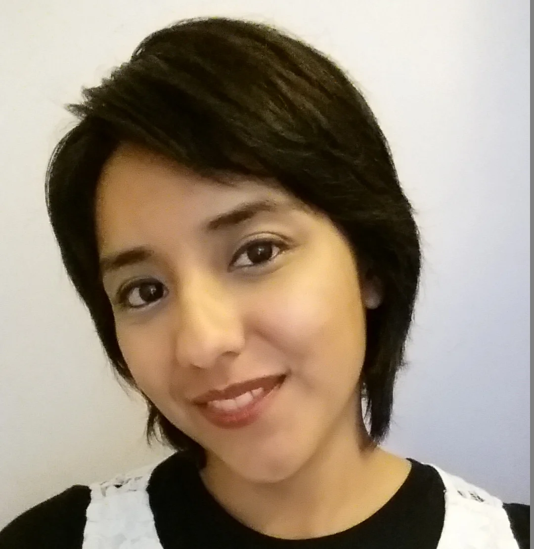 A person with short, dark hair smiles slightly at the camera. They are wearing a black top with a white lace detail and are posed in front of a plain light background.
