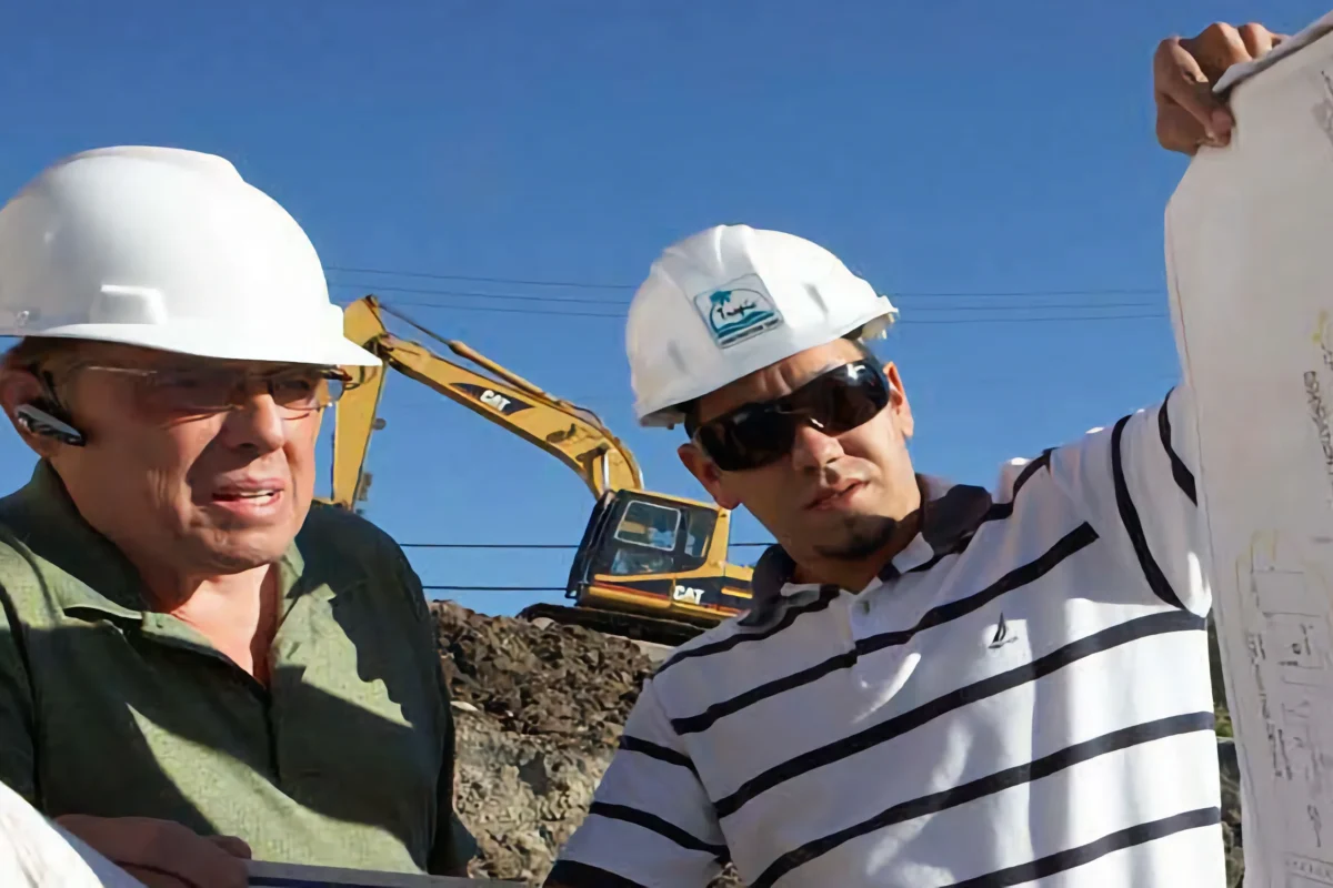 Two men wearing hard hats review construction plans at a worksite, with an excavator and dirt mound in the background under a clear blue sky.