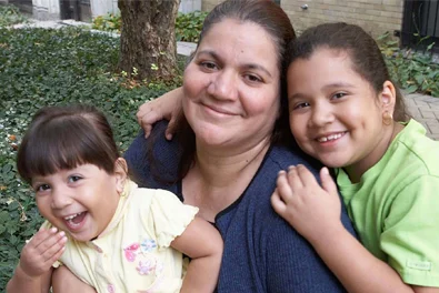 A woman sits outdoors with two smiling young girls, one on each side, in front of greenery and a brick building.