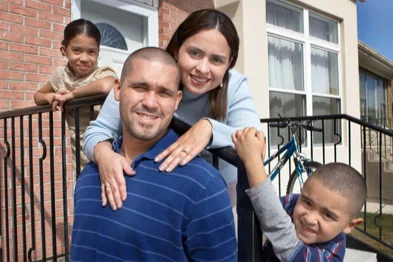A family of four poses and smiles in front of a house, with a man and woman in the center and two children, one girl and one boy, on either side.