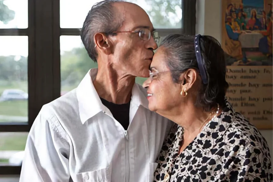 An elderly man kisses an elderly woman on the forehead while standing indoors near a window.