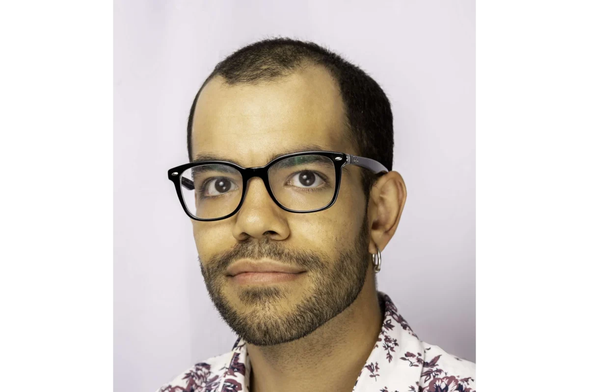 A person with short dark hair, glasses, a beard, and an earring is facing the camera against a light background, wearing a patterned shirt.