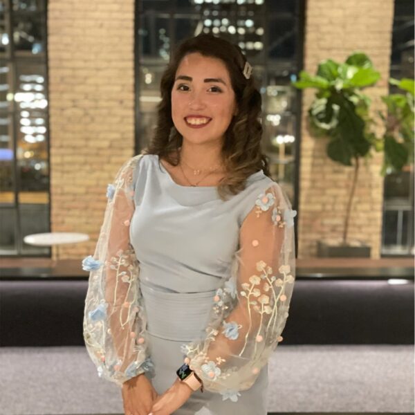 A woman in a light blue dress with floral sheer sleeves stands indoors, smiling at the camera. There are brick walls and large windows in the background.