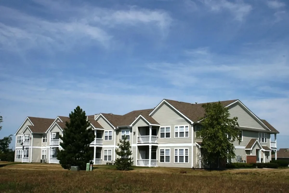Two-story apartment building with light gray siding and white trim, surrounded by grass and a few trees under a blue sky.