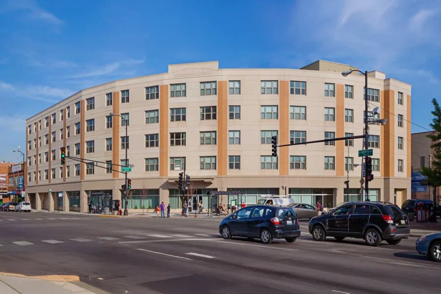 A modern four-story apartment building with beige and white exterior, large windows, and street-level shops at a busy intersection with cars and pedestrians.