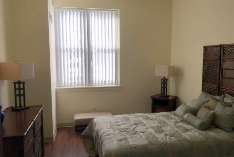 A bedroom with a single window, a dresser, ottoman, side table, and bed with many pillows and a headboard.