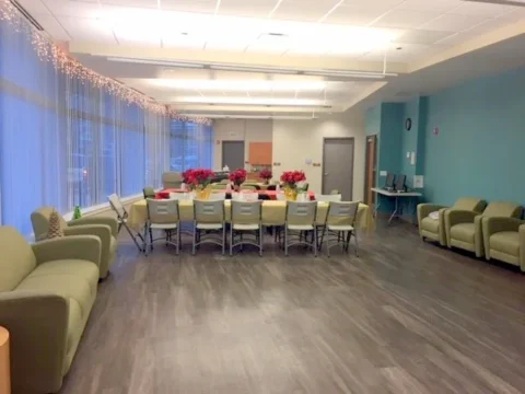 The community room has a large wooden floor, several cushioned arm chairs, and is set up with a couple tables, fold up chairs, and decorations for the holidays.