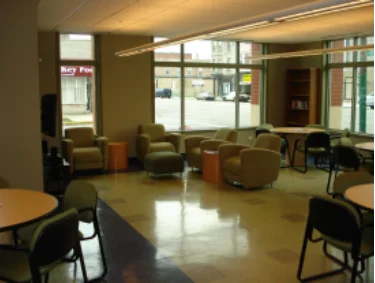 The community room has several cushioned armchairs, several round tables with chairs, and a large linoleum floor.