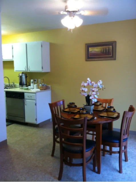 A small circular dining table with four chairs sits below a ceiling fan next to a galley kitchen.