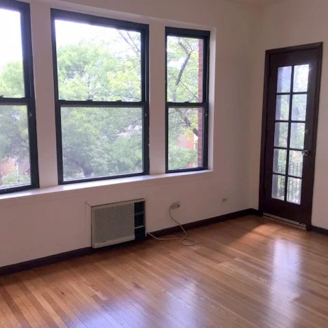 A wood-floored living room with several large windows and a door that leads to a balcony.
