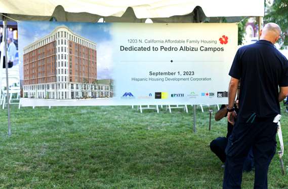 A banner under a tent displays a building rendering and text announcing the dedication of 1203 N. California Affordable Family Housing to Pedro Albizu Campos, dated September 1, 2023.
