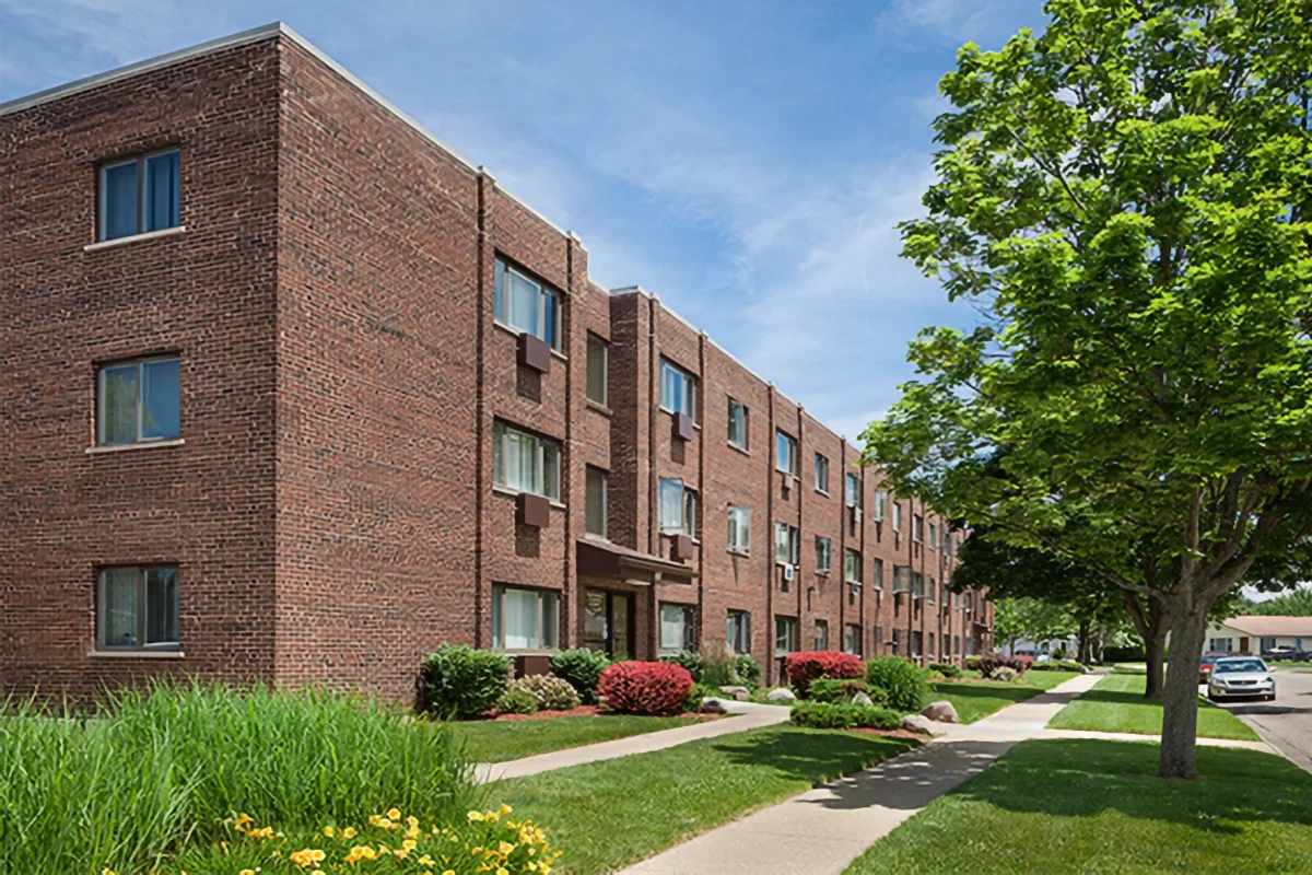 Three-story red brick apartment building with multiple windows, green landscaping, trees, and a sidewalk on a sunny day.