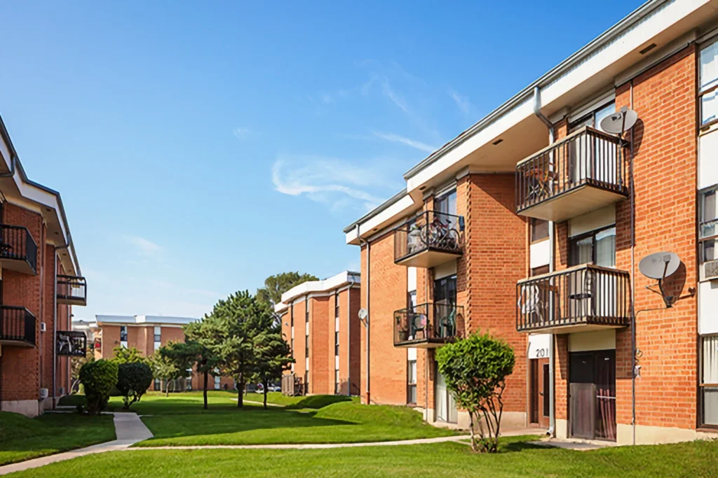 Red-brick apartment buildings with balconies and satellite dishes face a green lawn and walkway under a clear blue sky.