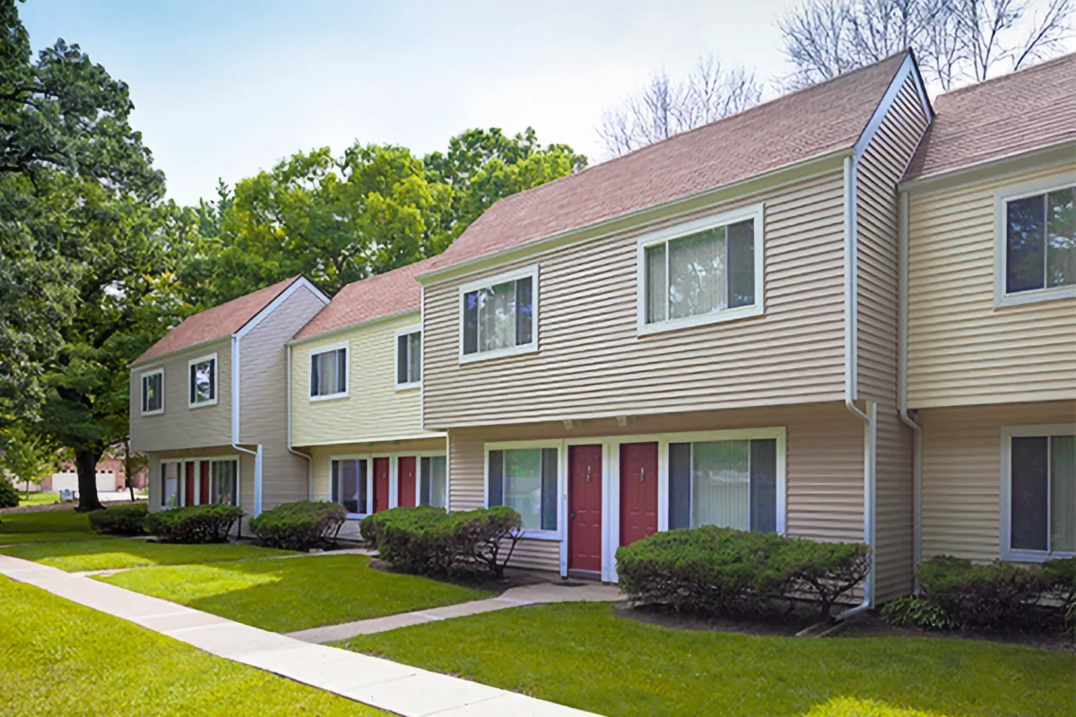 Row of beige townhouses with red doors, large windows, and well-kept shrubs along a sidewalk on a grassy lawn.