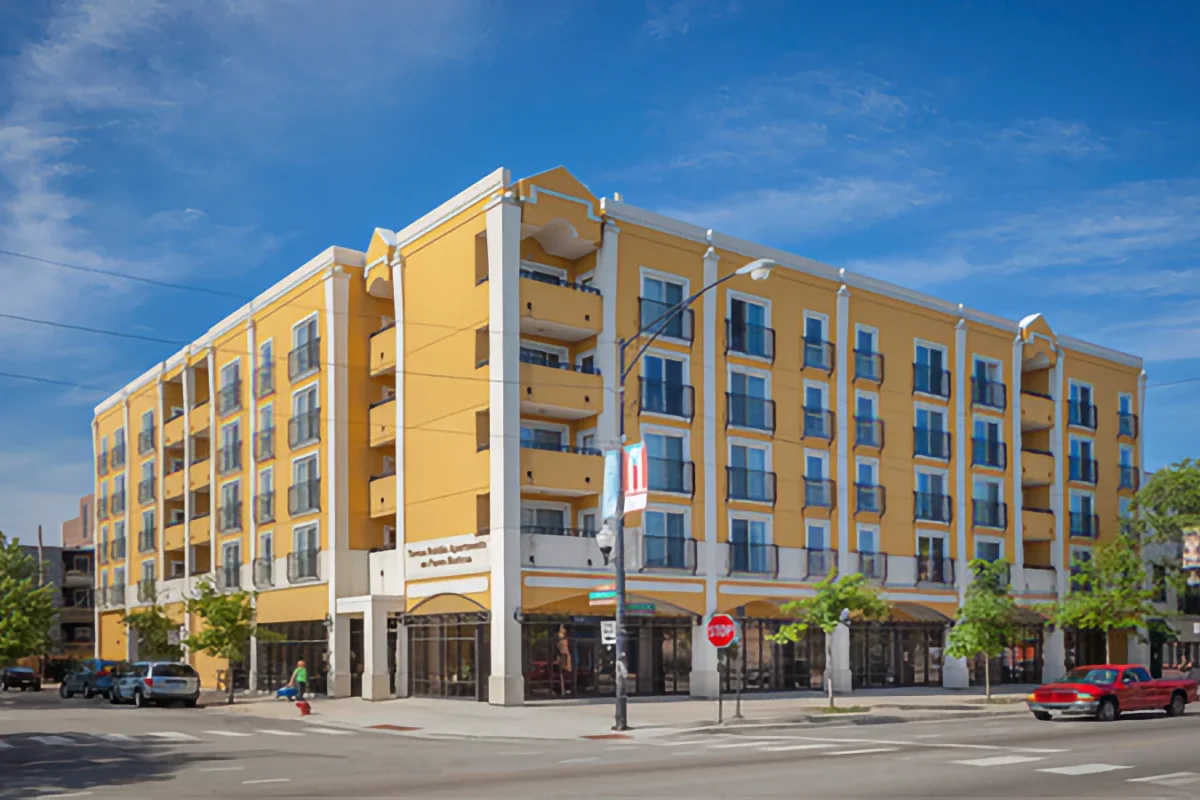 A four-story yellow apartment building with balconies sits on a street corner, with cars parked along the road and a clear blue sky above.