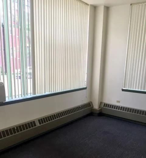 The corner of a living room showing two large windows, a blue carpet, and two baseboard heaters.