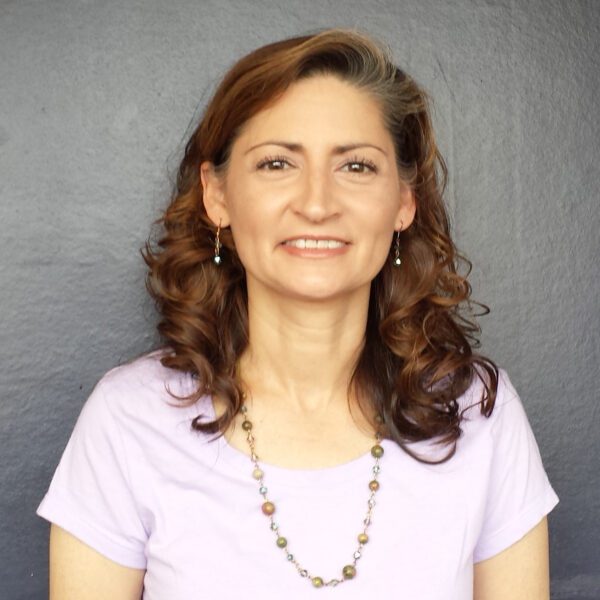 A woman with wavy brown hair wearing a light purple shirt, beaded necklace, and earrings stands in front of a plain dark background.