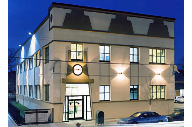 A modern, two-story building with large windows is lit up at night, featuring a glass entrance and a car parked outside.