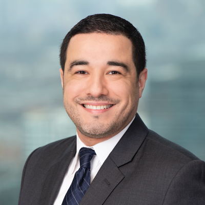 A man in a dark suit, white shirt, and striped tie smiles at the camera with a blurred office cityscape background.