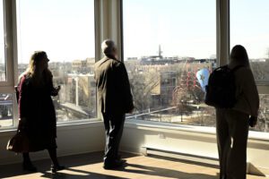 Three people stand inside a room with large windows, looking out at a cityscape in daylight.