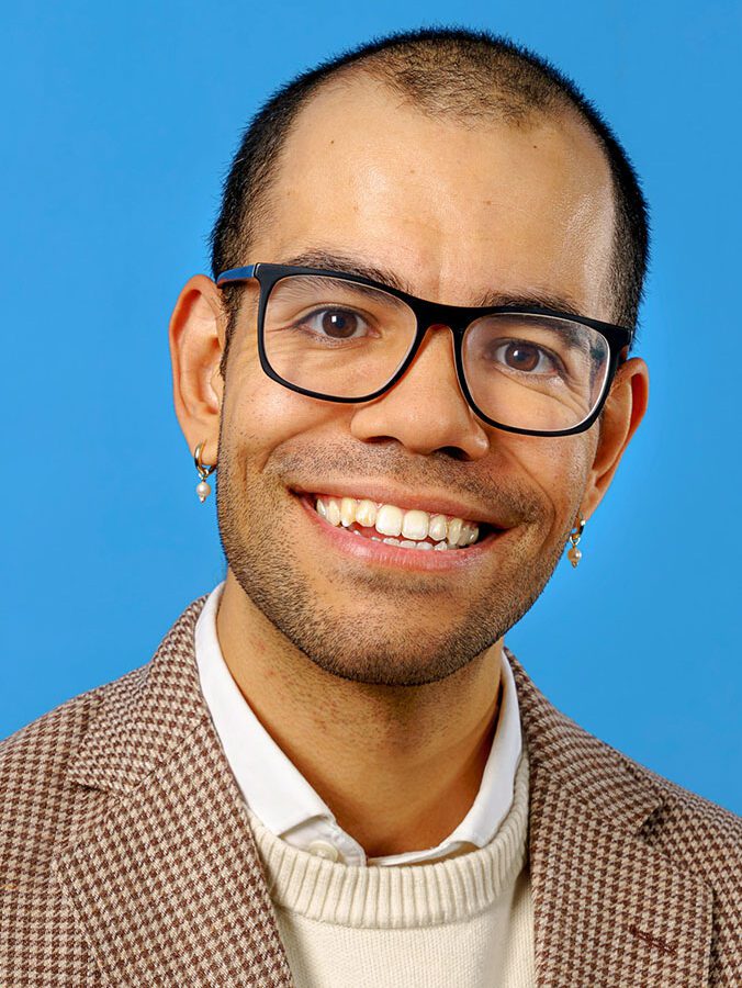 Smiling person with short hair, glasses, and earrings wearing a brown checkered blazer over a white sweater, posed in front of a blue background.